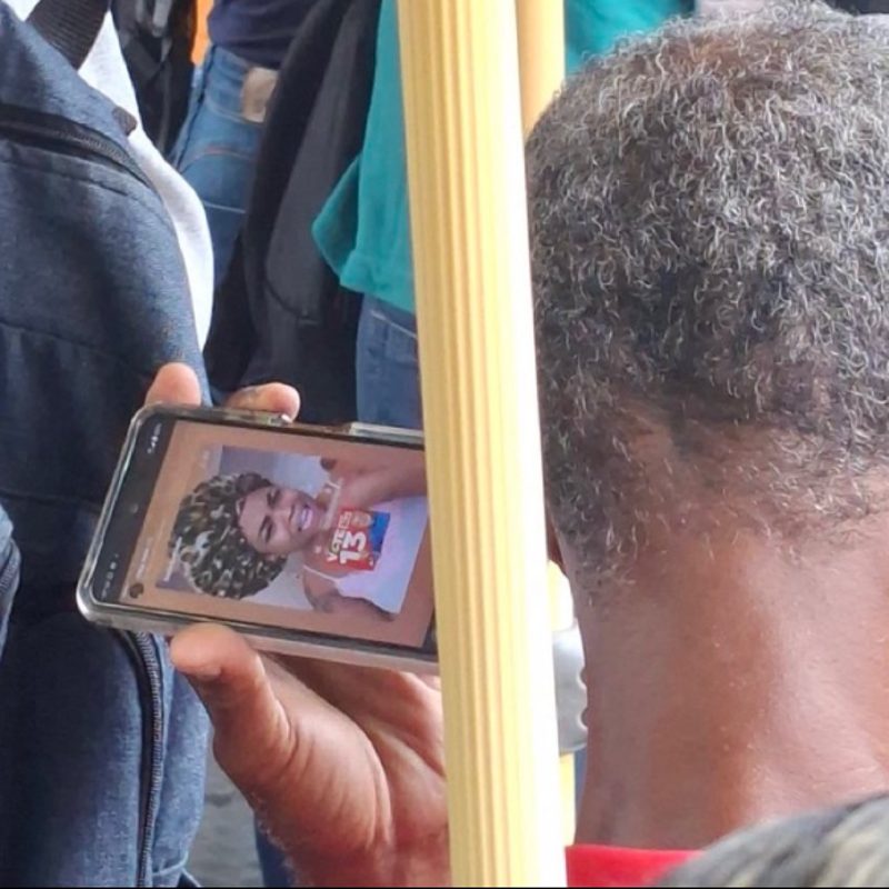 Homem segurando o celular com a imagem de uma pessoa de turbante e camiseta do 13, no ônibus